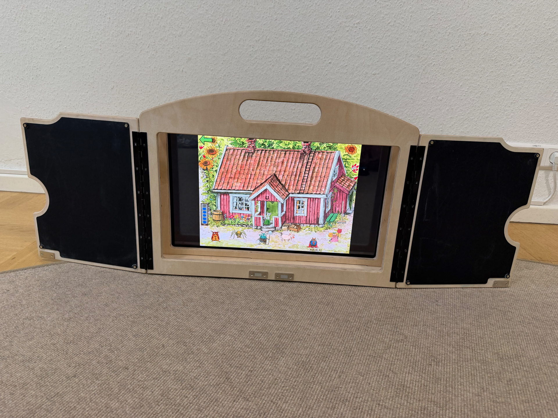 Das eKami liegt flach auf dem Boden; auf dem Bildschirm läuft ein Spiel mit einem roten Haus.