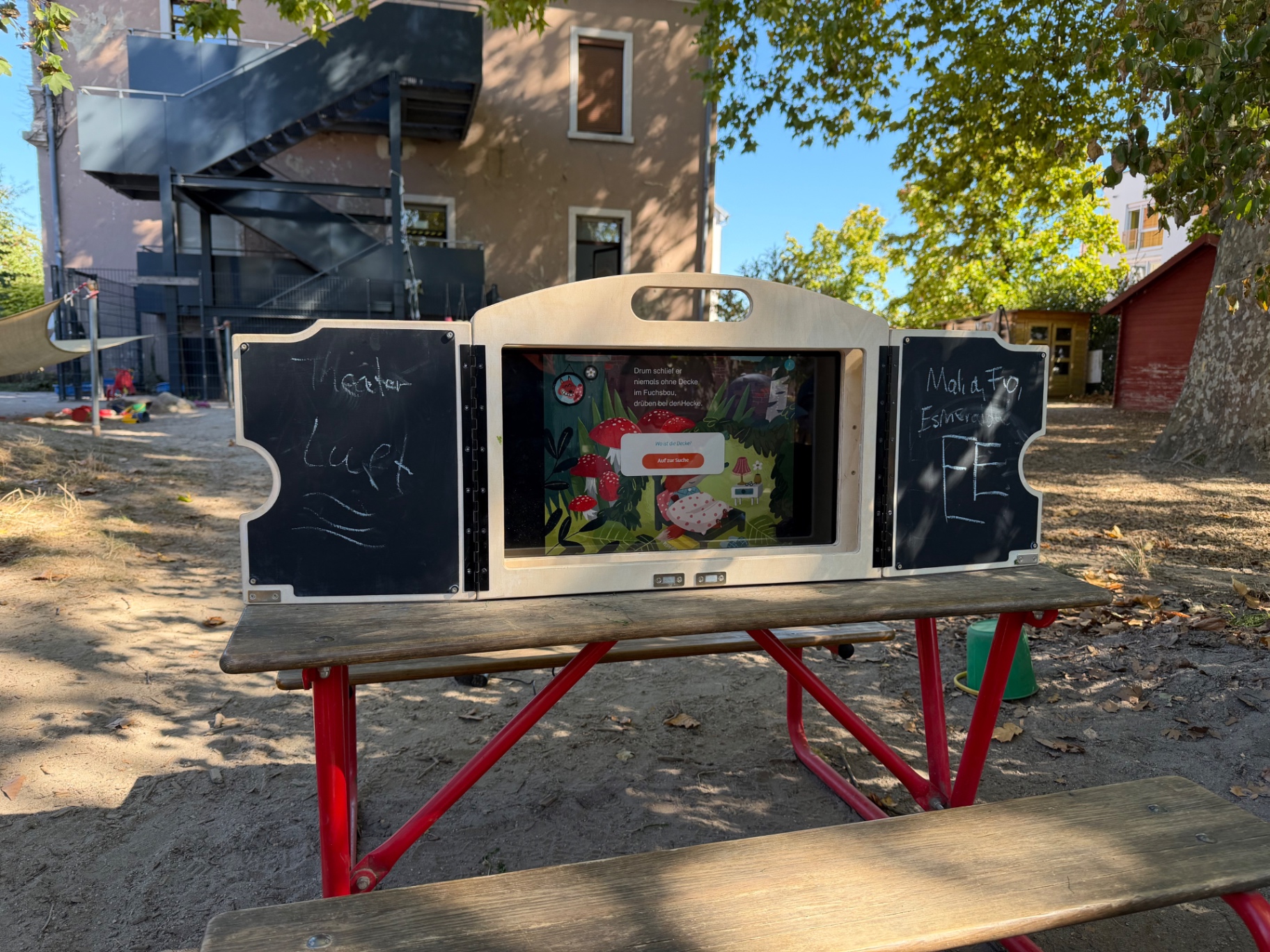 Das eKami mit Tafeln steht draußen auf einem Picknicktisch und zeigt eine Bilderbuchgeschichte mit Pilzen und Rotkäppchen.